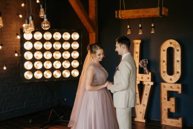 Bej takım elbiseli damat ve lavantalı gelin parlak stüdyoda dar bir gelinlik.