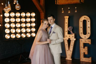 Bej takım elbiseli damat ve lavantalı gelin parlak stüdyoda dar bir gelinlik.