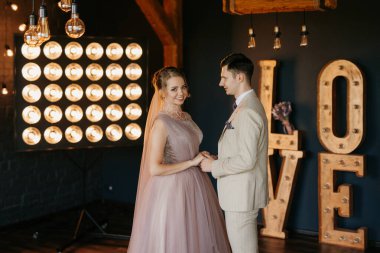 Bej takım elbiseli damat ve lavantalı gelin parlak stüdyoda dar bir gelinlik.