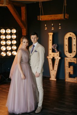 Bej takım elbiseli damat ve lavantalı gelin parlak stüdyoda dar bir gelinlik.