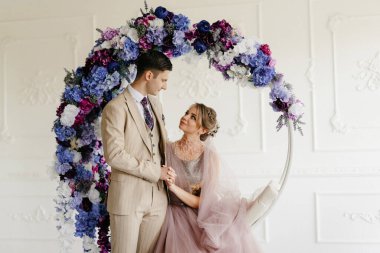 Bej takım elbiseli damat ve lavantalı gelin parlak stüdyoda dar bir gelinlik.