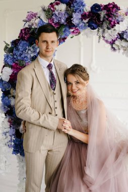 Bej takım elbiseli damat ve lavantalı gelin parlak stüdyoda dar bir gelinlik.