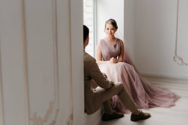 Bej takım elbiseli damat ve lavantalı gelin parlak stüdyoda dar bir gelinlik.