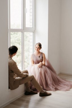 Bej takım elbiseli damat ve lavantalı gelin parlak stüdyoda dar bir gelinlik.