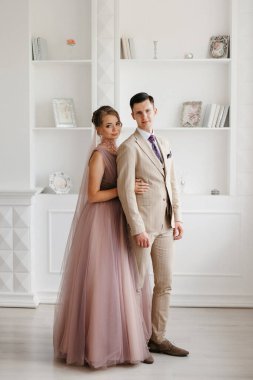 Bej takım elbiseli damat ve lavantalı gelin parlak stüdyoda dar bir gelinlik.