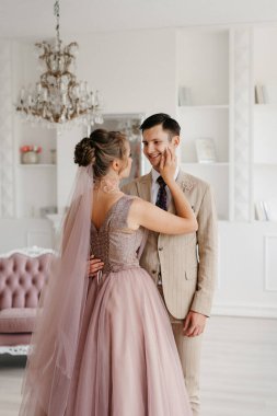 Bej takım elbiseli damat ve lavantalı gelin parlak stüdyoda dar bir gelinlik.