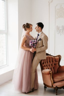 Bej takım elbiseli damat ve lavantalı gelin parlak stüdyoda dar bir gelinlik.