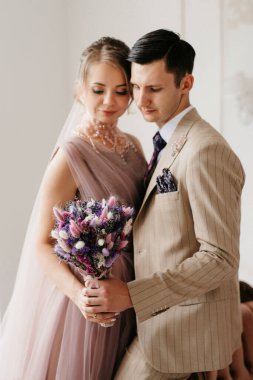 Bej takım elbiseli damat ve lavantalı gelin parlak stüdyoda dar bir gelinlik.