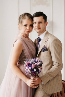 Bej takım elbiseli damat ve lavantalı gelin parlak stüdyoda dar bir gelinlik.