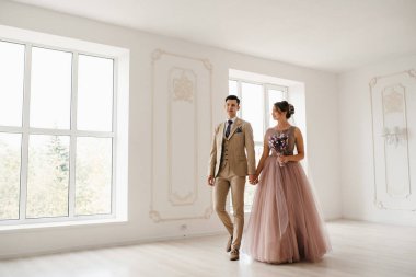 Bej takım elbiseli damat ve lavantalı gelin parlak stüdyoda dar bir gelinlik.