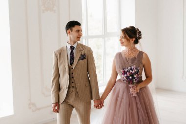 Bej takım elbiseli damat ve lavantalı gelin parlak stüdyoda dar bir gelinlik.