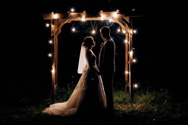 Gelin ve damat, sıcak bir ateş eşliğinde bir akşam düğün kemerine karşı.