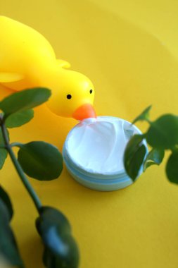 Kauçuk ördek ve yeşil yapraklı bir çerçevede sarı arka planda bir kavanoz krema.