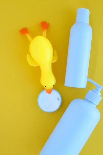 Sarı arka planda, vücut kreminin yanında duran plastik bir ördek var. Arka planda da bakım ürünü için mavi bir şişe var..