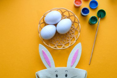 Süslü Paskalya yumurtaları olan hasır bir tabak sarı arka planda duruyor. Paskalya tavşanı ve yumurta boyası