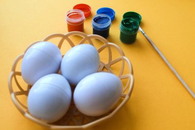 Süslü Paskalya yumurtaları olan hasır bir tabak sarı arka planda duruyor. Yumurta süslemek için boyalar