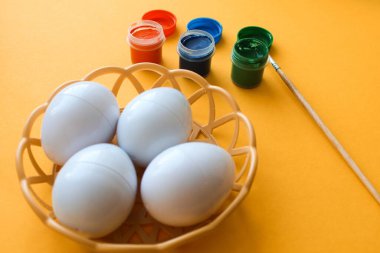 Süslü Paskalya yumurtaları olan hasır bir tabak sarı arka planda duruyor. Yumurta süslemek için boyalar