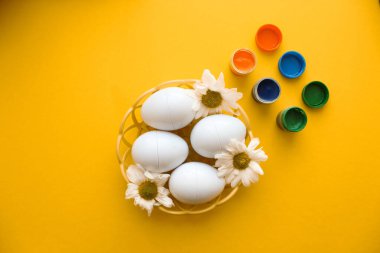 Süslü Paskalya yumurtaları olan hasır bir tabak sarı arka planda duruyor. Yumurta süslemek için boyalar