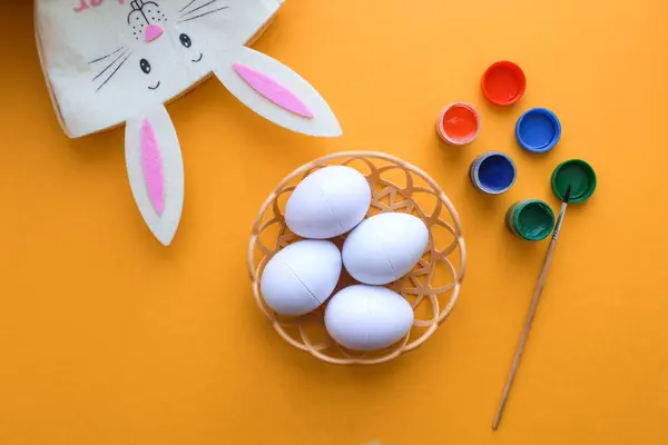 Süslü Paskalya yumurtaları olan hasır bir tabak sarı arka planda duruyor. Paskalya tavşanı ve yumurta boyası
