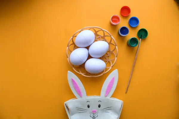 Süslü Paskalya yumurtaları olan hasır bir tabak sarı arka planda duruyor. Paskalya tavşanı ve yumurta boyası