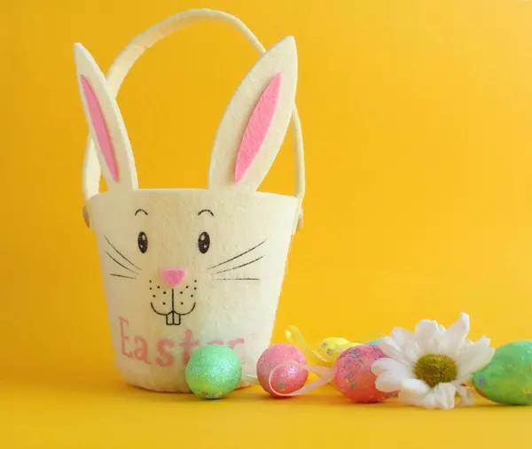 Sarı arka planda tavşan şeklinde bir yumurta sepeti ve Paskalya dekoratif yumurtaları var.