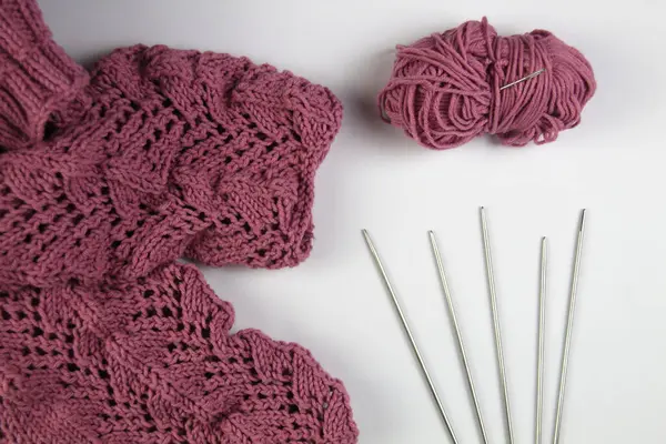 Pembe bir el yapımı süveter beyaz bir arka planda yatar. Örgü iğneleri ve iplik yumağı.