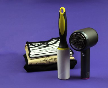 Katlanmış giysiler mor bir arka planda yatar. Ön plana bir askı makinesi ve bir giysi silindiri yerleştiriliyor..