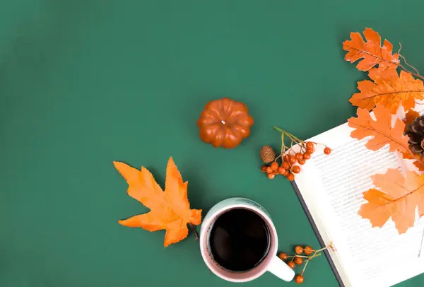 Yeşil arka planda bir kitap var, üzerinde akçaağaç yaprakları, aromatik kahve ve turuncu balkabağı var..