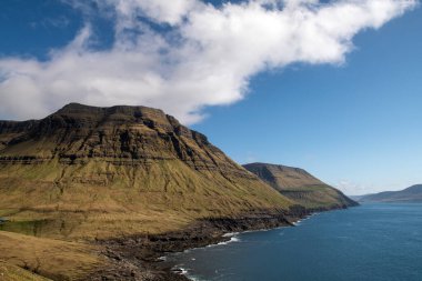 Faroe Adaları 'nda güzel bir deniz manzarası var. Mavi gökyüzüne karşı beyaz bulutlar ve baharda çimlerle kaplı dağlar..