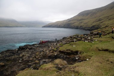 Turistler bulutlu bir günde Faroe Adaları 'ndaki Drangarnir sahillerinde pirzola teknesiyle alınıyorlar. Arkaplanda dağlar.