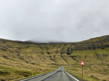 Faroese yolunda keskin dönüş işareti. Sisin dağların üzerinde süzüldüğü gün dolambaçlı bir yol.