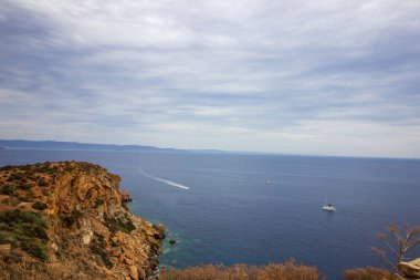 Sakin kıyı manzarası sakin mavi denizle buluşan kayalık uçurumların çarpıcı görüntüsü, sularda süzülen yelkenliler yumuşak bulutlu bir gökyüzü altında, Yüksek kalite fotoğraf