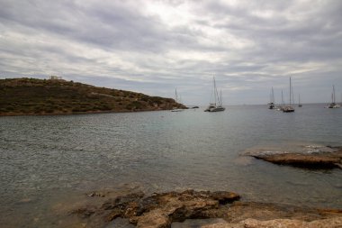 Sakin kıyı manzarası sakin mavi denizle buluşan kayalık uçurumların çarpıcı görüntüsü, sularda süzülen yelkenliler yumuşak bulutlu bir gökyüzü altında, Yüksek kalite fotoğraf