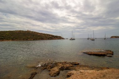 Sakin kıyı manzarası sakin mavi denizle buluşan kayalık uçurumların çarpıcı görüntüsü, sularda süzülen yelkenliler yumuşak bulutlu bir gökyüzü altında, Yüksek kalite fotoğraf