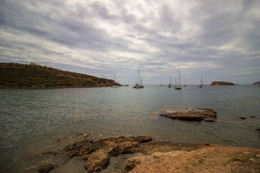 Sakin kıyı manzarası sakin mavi denizle buluşan kayalık uçurumların çarpıcı görüntüsü, sularda süzülen yelkenliler yumuşak bulutlu bir gökyüzü altında, Yüksek kalite fotoğraf