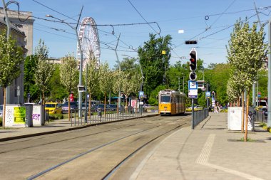 Macaristan 'ın Budapeşte şehrinin arka planında trafik ışıkları ve dönme dolabı olan ağaçlık bir sokaktan geçen sarı tramvay. Kentsel ulaşım ve şehir manzarası. 