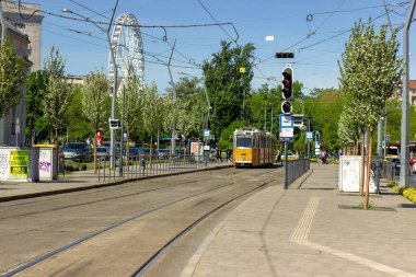 Macaristan 'ın Budapeşte şehrinin arka planında trafik ışıkları ve dönme dolabı olan ağaçlık bir sokaktan geçen sarı tramvay. Kentsel ulaşım ve şehir manzarası. 