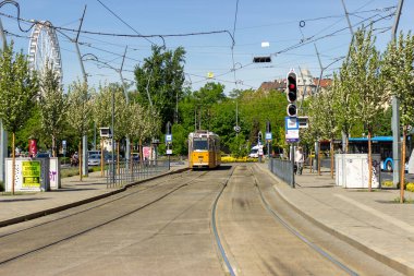 Macaristan 'ın Budapeşte şehrinin arka planında trafik ışıkları ve dönme dolabı olan ağaçlık bir sokaktan geçen sarı tramvay. Kentsel ulaşım ve şehir manzarası. 