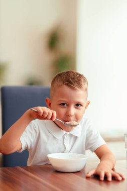 Evde kahvaltı yapan, suyla yulaf lapası yiyen 5 yaşında tatlı bir çocuk. Doğal ışık, sağlıklı yiyecekler, yemek masasındaki çocuk, sıcak aile anı..