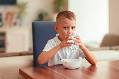 Evde kahvaltı yapan, suyla yulaf lapası yiyen 5 yaşında tatlı bir çocuk. Doğal ışık, sağlıklı yiyecekler, yemek masasındaki çocuk, sıcak aile anı..