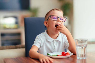 Sevimli 5 yaşında gözlüklü bir çocuk masada oturmuş domates ve dolmalık biber gibi taze sebze yiyor. O tatlı bir ifadeyle yiyeceğe bakar. Sağlıklı beslenme, çocuk beslenmesi ve ev hayatı..