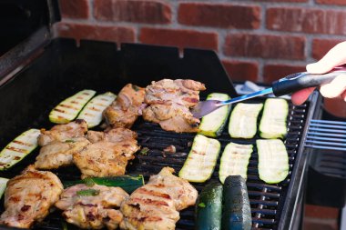 Hand holding piece of grilled meat while grilling chicken and zukini vegetables on outdoor barbecue grill close up photo. Food background texture. Weekend, picnic or party concept.