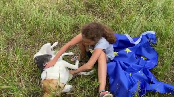 4K小女孩和她的狗在草坪上的欧洲大旗上放松 女孩坐在草地上 轻抚着狗 摄像机的运动 — 图库视频影像