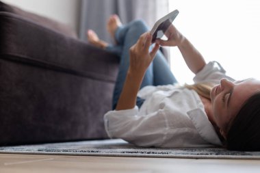 Genç bir kadın elinde cep telefonuyla evde halıya uzanıyor ve dinleniyor. Seçici odak