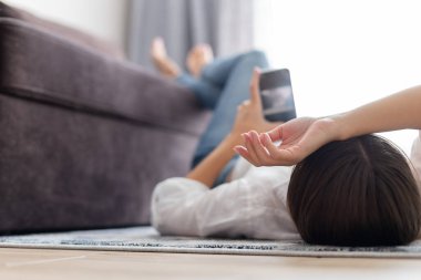 Genç bir kadın evde halıya uzanmış, rahatlıyor ve cep telefonu kullanıyor. Seçici odak
