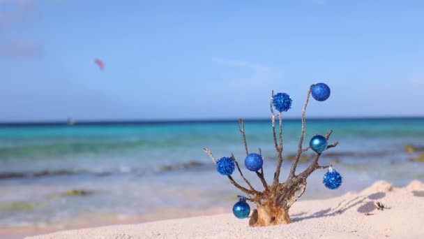 青いクリスマスボールが後ろに青い海がある砂浜に立っている死んだサンゴで飾られ カイトサーファーが離れています 4K映像 — ストック動画