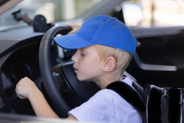 Little boy in a blue cap and with school bag sitting on the driver seat in a car. Little boy wants to start the car