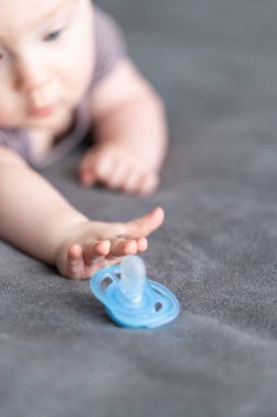 Gri battaniyeye uzanmış bebek, mavi emziğe ulaşmak için geriniyor. Seçici odak, yakın plan