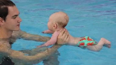 Bebek kapalı havuzda yüzüyor. Koç baba yeni doğmuş bebeği tutuyor, yüzme havuzunda mavi suda yüzmeyi öğretiyor. Koç yardımıyla yüzmeye çalışan küçük bir çocuk. 4k görüntü.