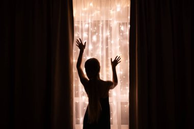 Karanlık silueti olan bir kadın Noel aydınlatmasıyla pencerenin yanında duruyor.
